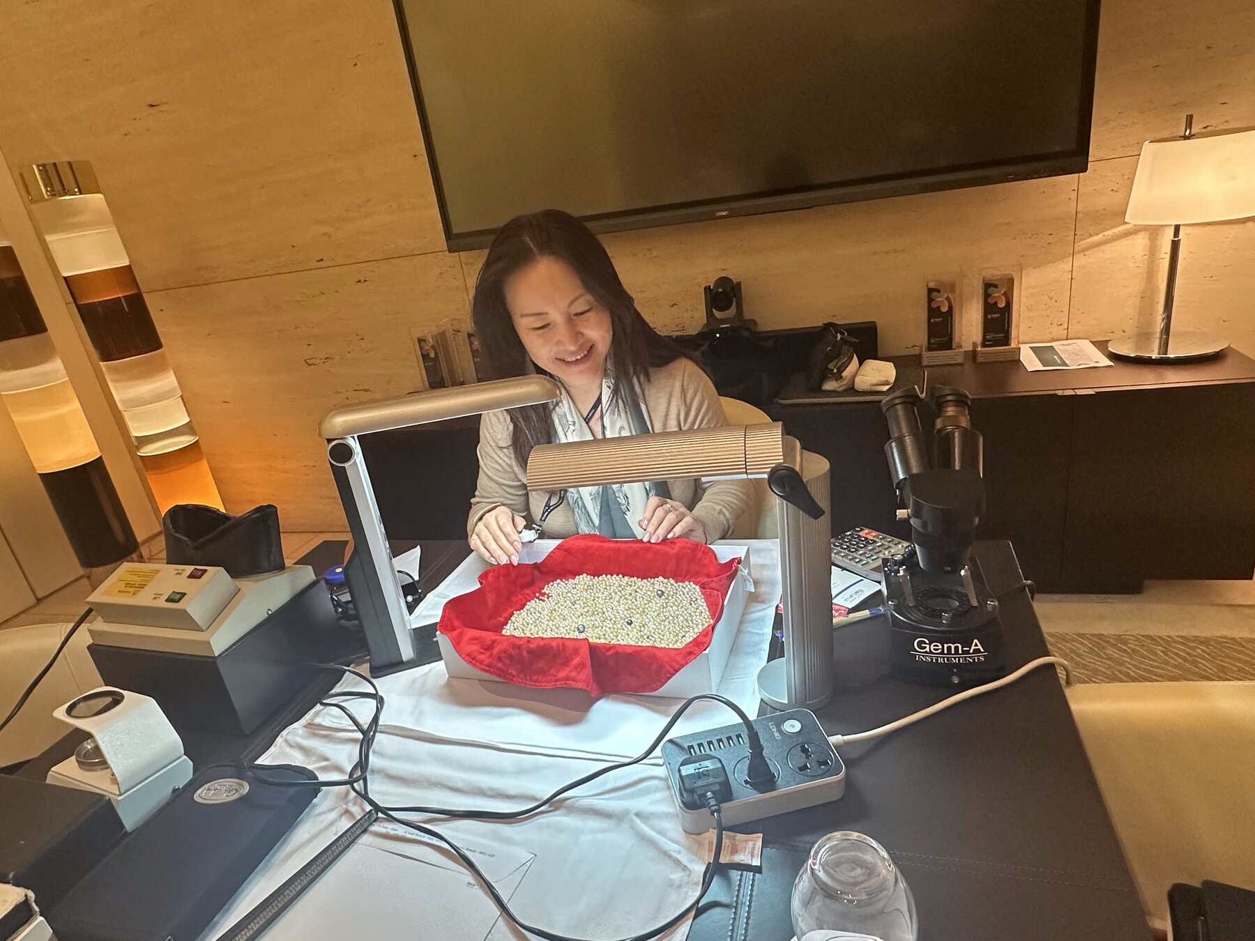Kehan Li seated at her microscope station, smiling, with a tray of pearls on red velvet before her.